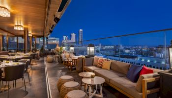 This image shows a stylish rooftop lounge with sofas, tables, and stools, overlooking a cityscape at night with buildings in the background.