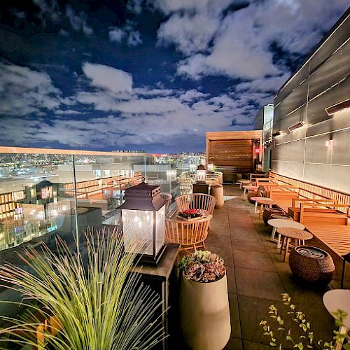 A rooftop lounge area with modern furniture, plants, and lanterns, offering a stunning cityscape view under a partly cloudy night sky.