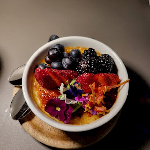 A bowl of dessert topped with strawberries, blueberries, blackberries, and edible flowers, placed on a table with a spoon beside it.