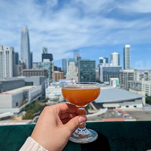 A hand holds a cocktail glass against a backdrop of a city skyline with tall buildings and a partly cloudy sky.