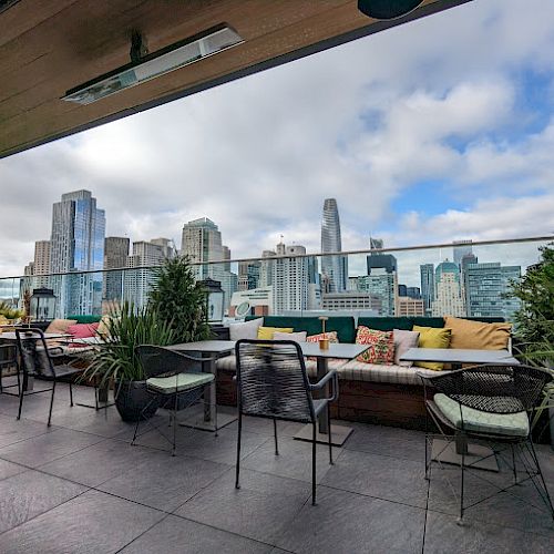 A rooftop patio with chairs and tables, surrounded by greenery, overlooks a modern city skyline against a partly cloudy sky.