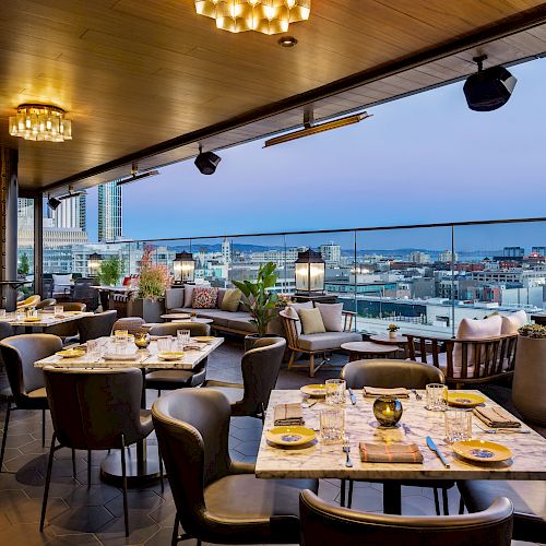 A rooftop restaurant with city views, featuring elegantly set tables, cushioned seating, warm lighting fixtures, and a modern, inviting ambiance.