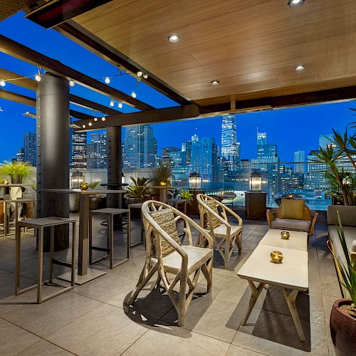 A stylish rooftop lounge with city skyline views, wooden ceiling, modern furniture, potted plants, and ambient lighting during the evening.